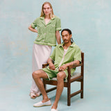Male model with dreadlocks and female model with long blonde hair posing together with the male model seated wearing the 'Standard Issue' Cabana (green) half-zipped with a white, mesh tanktop underneath, and matching 'Standard Issue' green shorts and white slippers while the female model stands behind him to the side with her hand on the chair as she wears the 'Rick Rack' Cabana ~ Crop Cut (Green) with a white silk full-length dress and white slippers, all shot on a light blue background.
