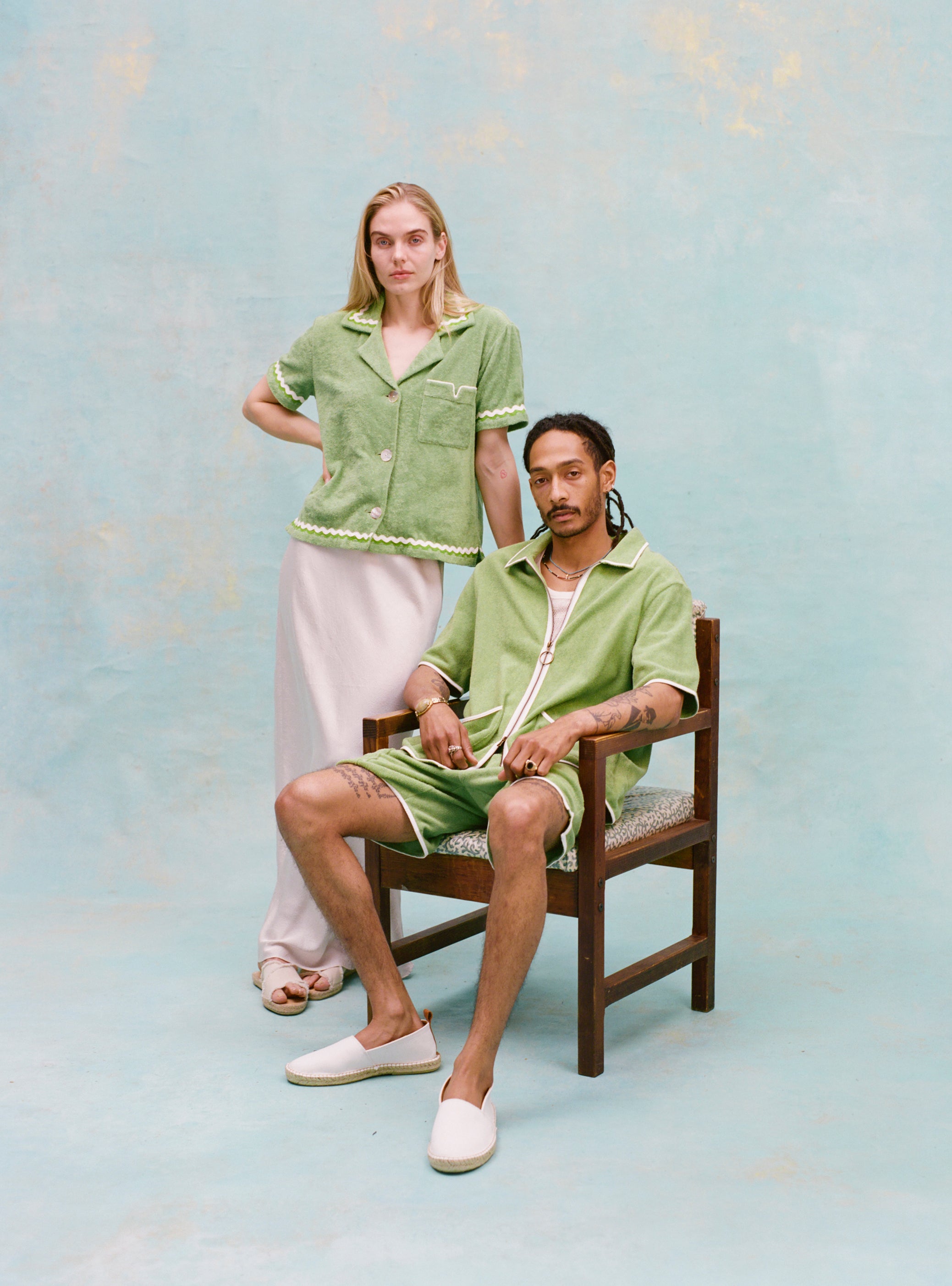 Male model with dreadlocks and female model with long blonde hair posing together with the male model seated wearing the 'Standard Issue' Cabana (green) half-zipped with a white, mesh tanktop underneath, and matching 'Standard Issue' green shorts and white slippers while the female model stands behind him to the side with her hand on the chair as she wears the 'Rick Rack' Cabana ~ Crop Cut (Green) with a white silk full-length dress and white slippers, all shot on a light blue background.