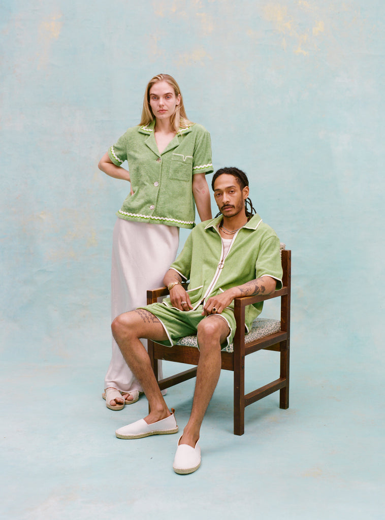 Male model with dreadlocks and female model with long blonde hair posing together with the male model seated wearing the 'Standard Issue' Cabana (green) half-zipped with a white, mesh tanktop underneath, and matching 'Standard Issue' green shorts and white slippers while the female model stands behind him to the side with her hand on the chair as she wears the 'Rick Rack' Cabana ~ Crop Cut (Green) with a white silk full-length dress and white slippers, all shot on a light blue background.