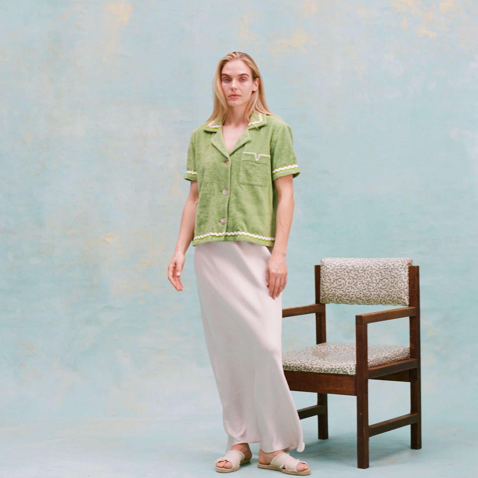 Female model with long blonde hair wearing the 'Rick Rack' Cabana ~Crop Cut (green) buttoned with full-length silk white dress and white slippers, with a wooden chair with white cushions in the background, all shot on a textured light blue background.