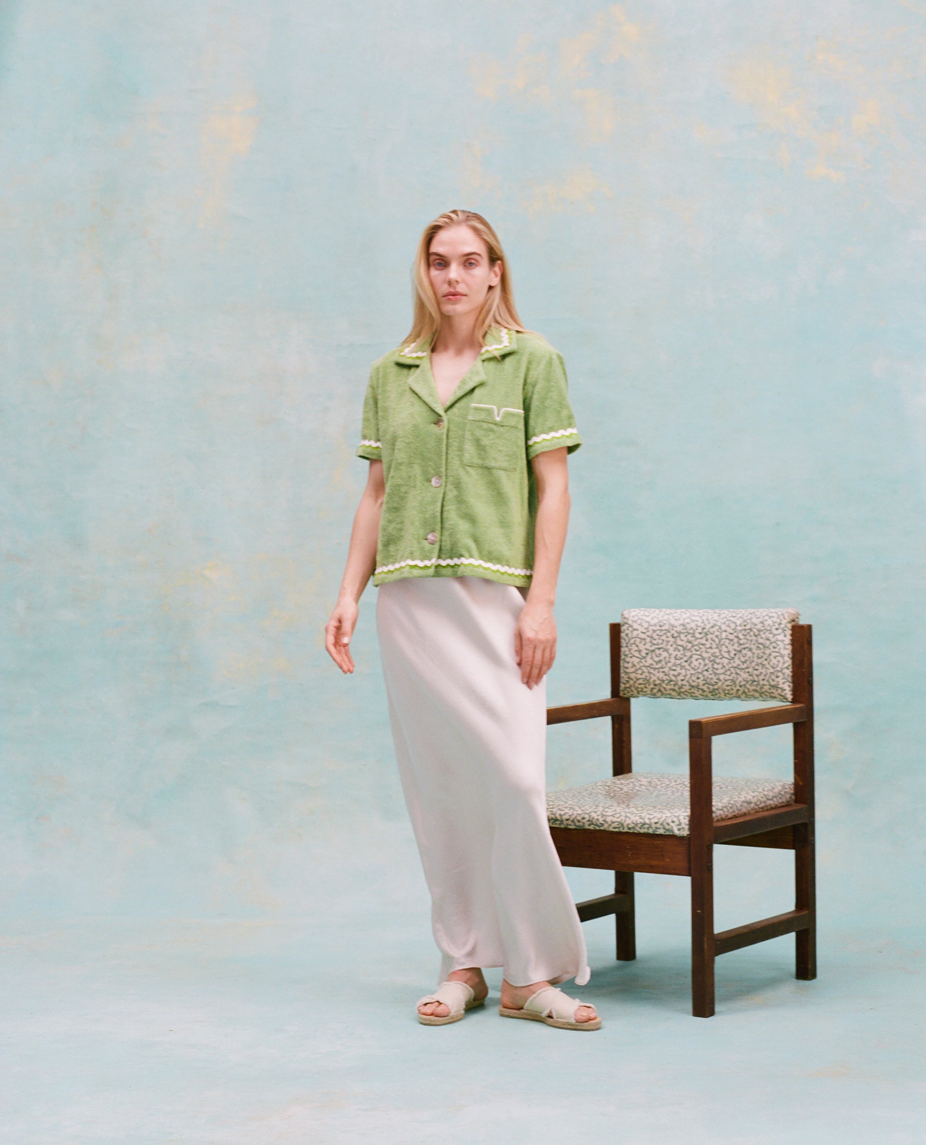 Female model with long blonde hair wearing the 'Rick Rack' Cabana ~Crop Cut (green) buttoned with full-length silk white dress and white slippers, with a wooden chair with white cushions in the background, all shot on a textured light blue background.