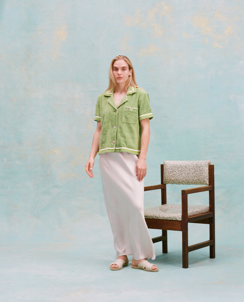 Female model with long blonde hair wearing the 'Rick Rack' Cabana ~Crop Cut (green) buttoned with full-length silk white dress and white slippers, with a wooden chair with white cushions in the background, all shot on a textured light blue background.