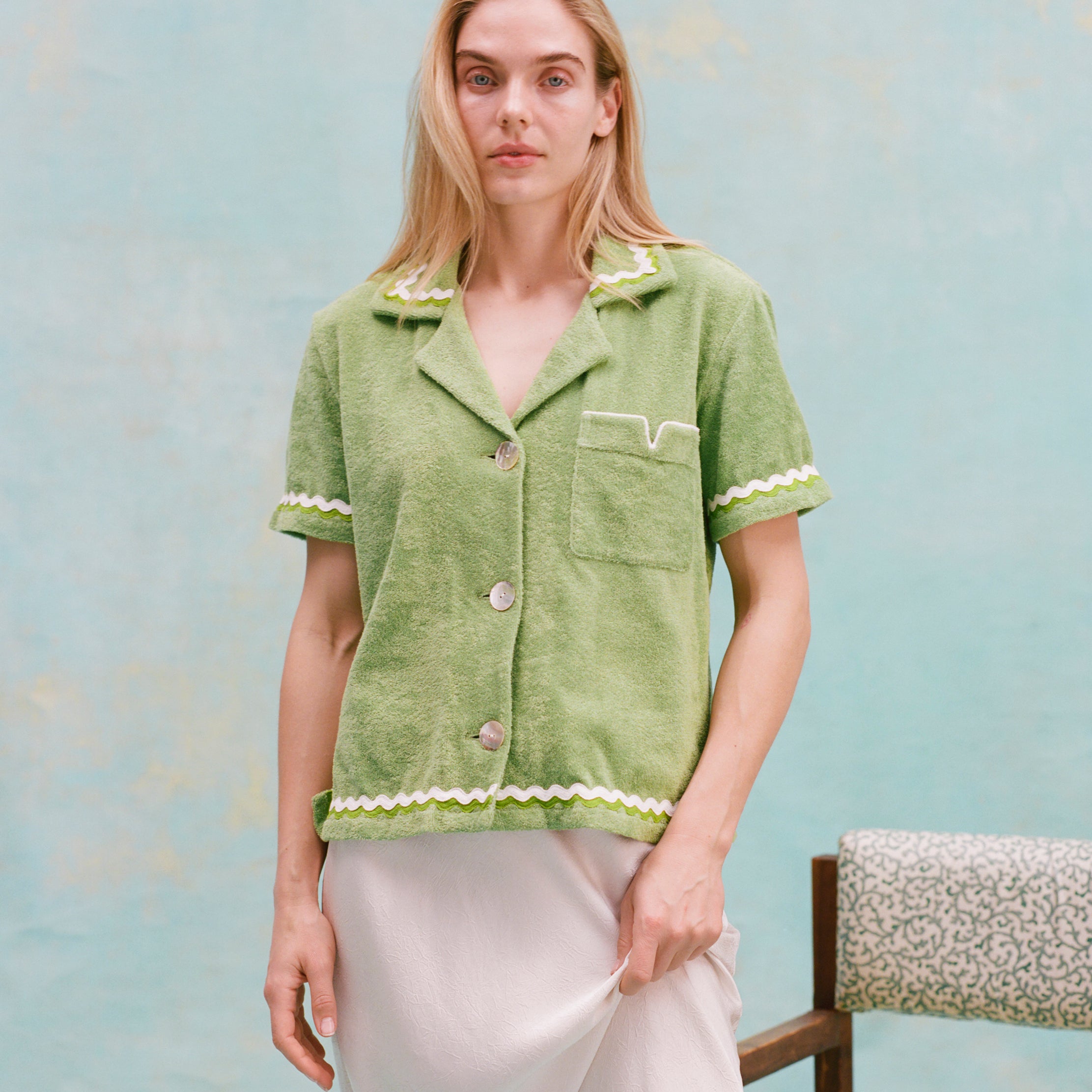 Female model with long blonde hair wearing the 'Rick Rack' Cabana ~ Crop Cut (Green) buttoned with a white full-length silk dress, all shot on a textured light blue background.