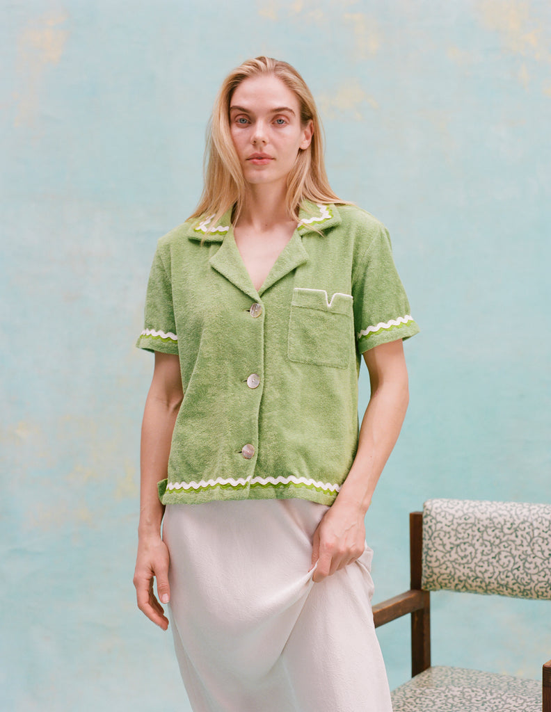 Female model with long blonde hair wearing the 'Rick Rack' Cabana ~ Crop Cut (Green) buttoned with a white full-length silk dress, all shot on a textured light blue background.