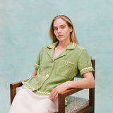 Female model with long blonde hair seated wearing the 'Rick Rack' Cabana ~ Crop Cut (green) buttoned with a full-length white silk dress, shot on a textured light blue background.