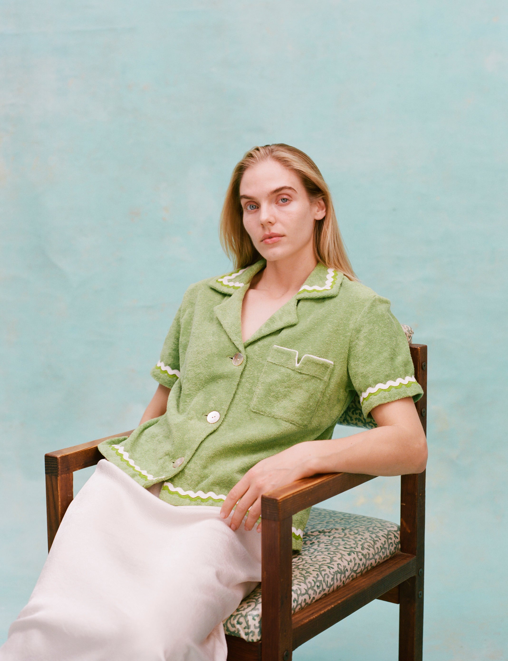 Female model with long blonde hair seated wearing the 'Rick Rack' Cabana ~ Crop Cut (green) buttoned with a full-length white silk dress, shot on a textured light blue background.
