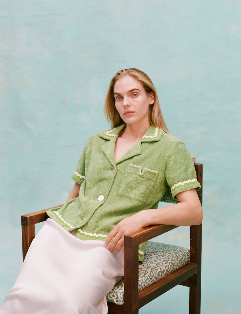 Female model with long blonde hair seated wearing the 'Rick Rack' Cabana ~ Crop Cut (green) buttoned with a full-length white silk dress, shot on a textured light blue background.