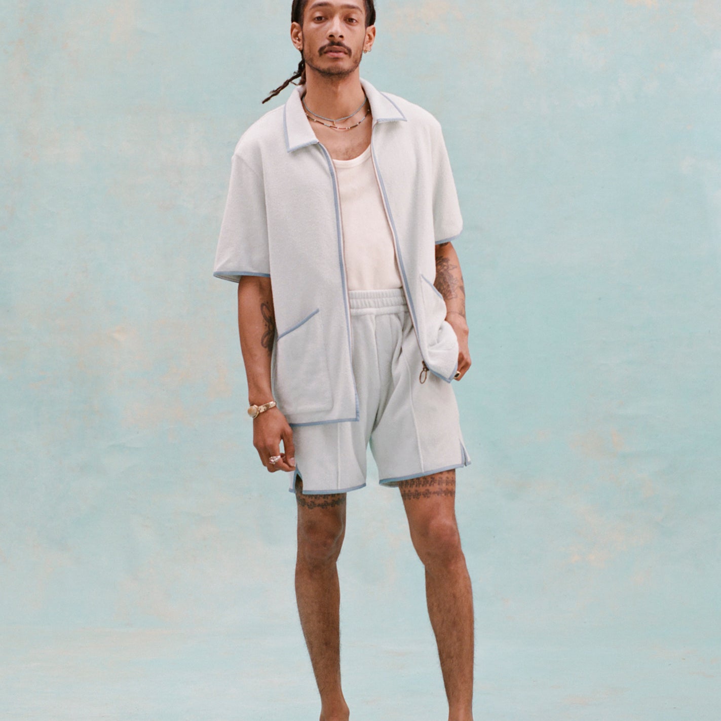 Male model with dreadlocks wearing the 'Standard Issue' Cabana (Sky Blue) fully unzipped with a white tank top underneath that is tucked into matching 'standard issue' shorts in Sky Blue with white slippers, all shot on a light blue background.