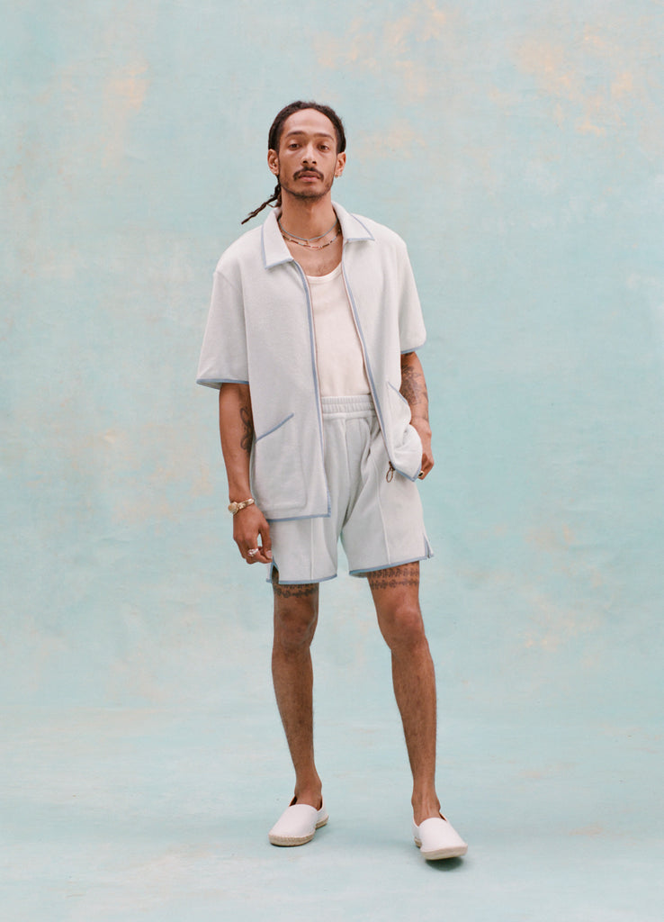 Male model with dreadlocks wearing the 'Standard Issue' Cabana (Sky Blue) fully unzipped with a white tank top underneath that is tucked into matching 'standard issue' shorts in Sky Blue with white slippers, all shot on a light blue background.