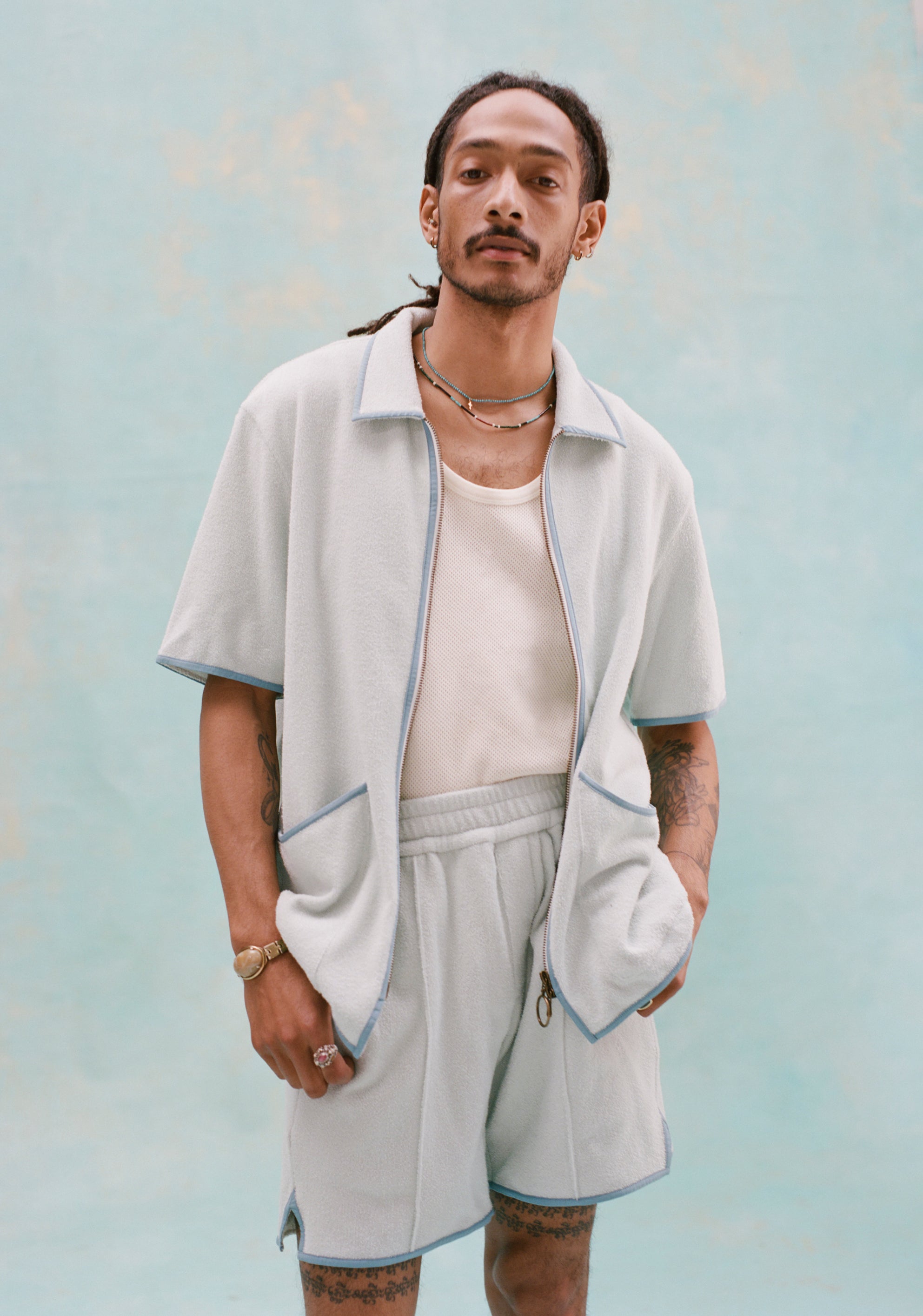 Male model with dreadlocks wearing a 'Standard Issue' Cabana (Sky Blue) that is fully unzipped, a white tank top underneath it tucked into matching 'Standard Issue' Sky blue shorts, all shot on a light blue background.