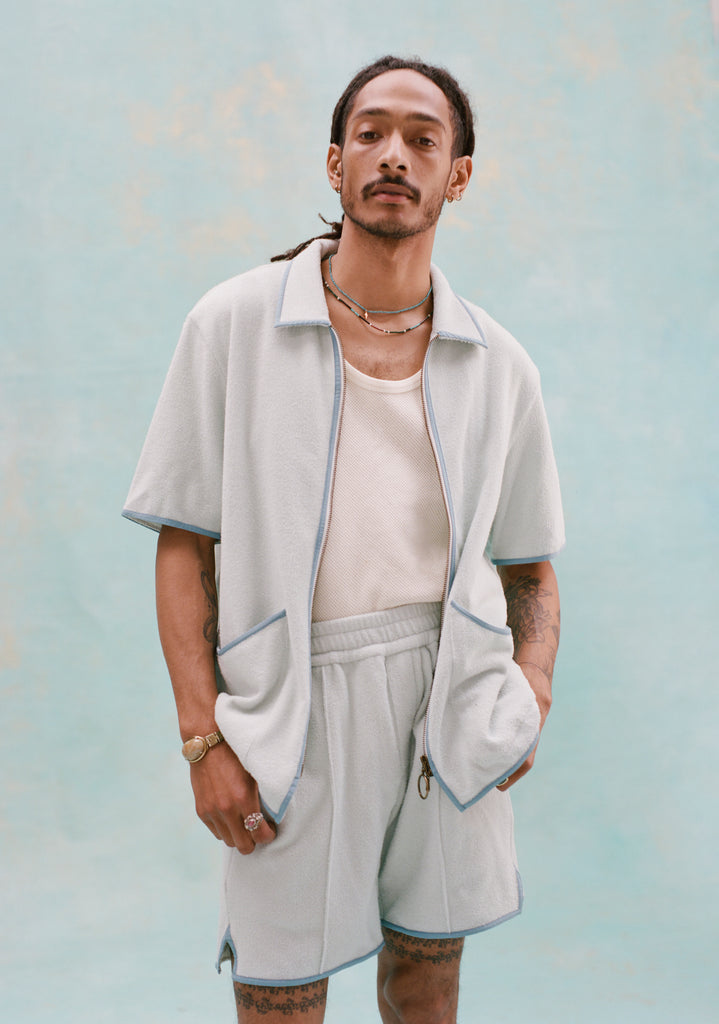 Male model with dreadlocks wearing a 'Standard Issue' Cabana (Sky Blue) that is fully unzipped, a white tank top underneath it tucked into matching 'Standard Issue' Sky blue shorts, all shot on a light blue background.