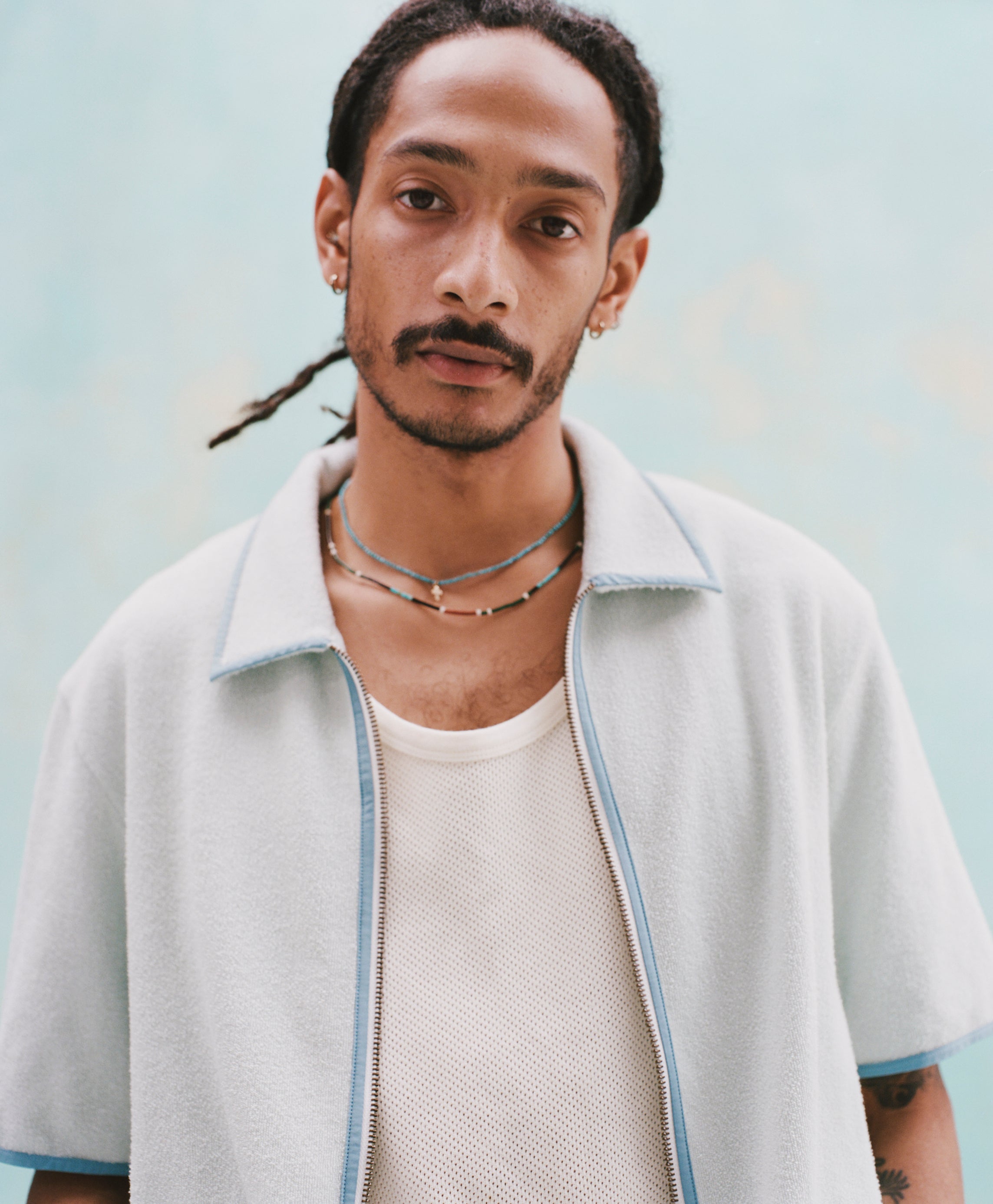 Male mode with dreadlocks wearing the 'Standard Issue' Cabana (Sky Blue) fully unzipped with a white tank top underneath, shot against a light blue background.
