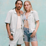 Male model with dreadlocks and female model with long blonde hair posing together with the male model wearing the 'Standard Issue' Cabana (Sky Blue) fully unzipped with a white tanktop underneath tucked into matching 'Standard Issue' shorts in sky blue while the female model, with her right hand over the shoulder of the male model, is wearing a 'Rick Rack' Cabana ~ Crop Cut (Sky Blue) unbuttoned with a light blue bikini top, and jean shorts, shot on a light blue background.