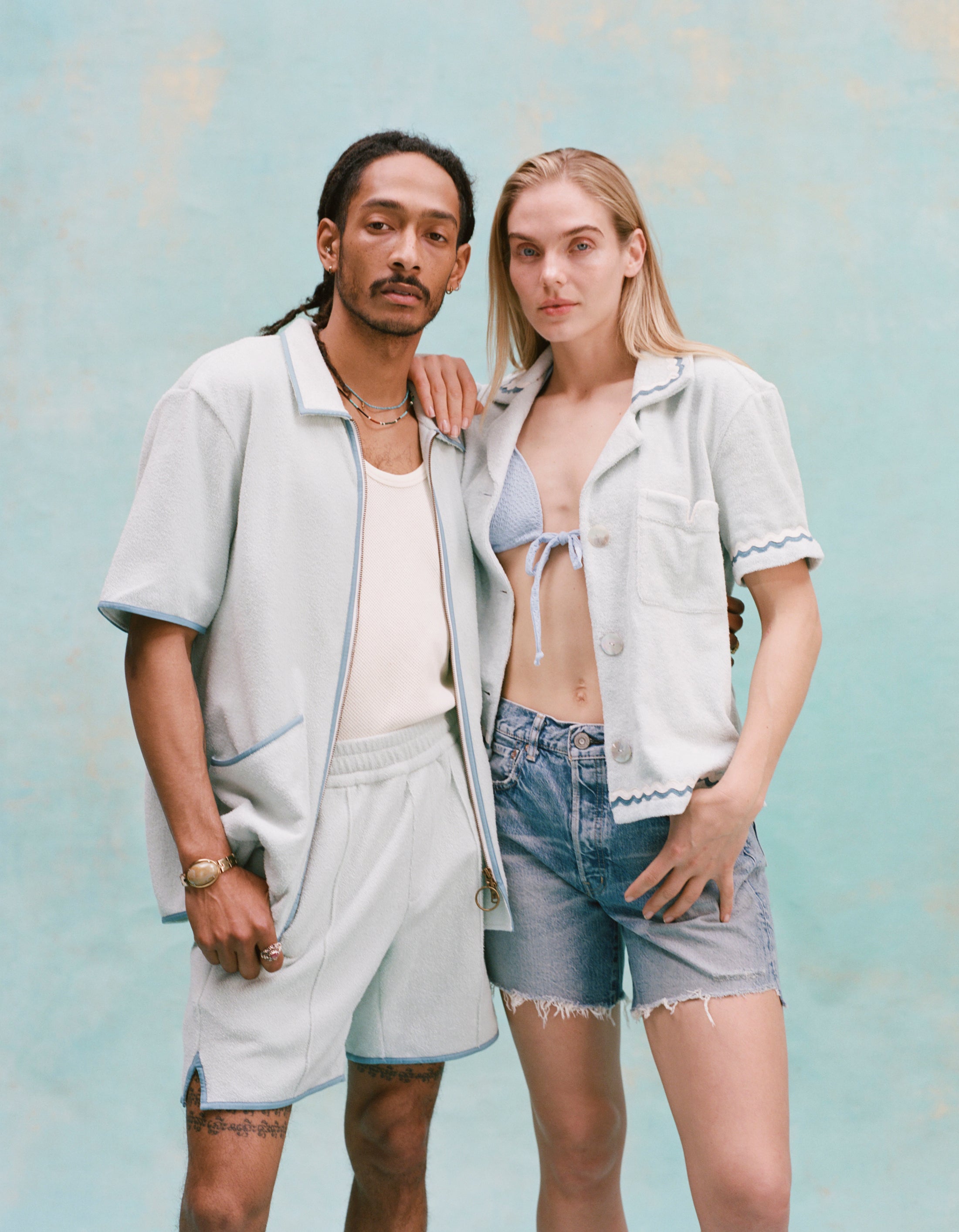 Male model with dreadlocks and female model with long blonde hair posing together with the male model wearing the 'Standard Issue' Cabana (Sky Blue) fully unzipped with a white tanktop underneath tucked into matching 'Standard Issue' shorts in sky blue while the female model, with her right hand over the shoulder of the male model, is wearing a 'Rick Rack' Cabana ~ Crop Cut (Sky Blue) unbuttoned with a light blue bikini top, and jean shorts, shot on a light blue background.