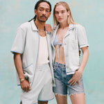 Male model with dreadlocks and female model with long blonde hair posing together with the male model wearing the 'Standard Issue' Cabana (Sky Blue) fully unzipped with a white tanktop underneath tucked into matching 'Standard Issue' shorts in sky blue while the female model, with her right hand over the shoulder of the male model, is wearing a 'Rick Rack' Cabana ~ Crop Cut (Sky Blue) unbuttoned with a light blue bikini top, and jean shorts, shot on a light blue background.