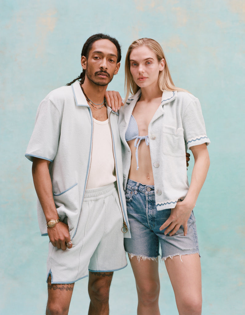 Male model with dreadlocks and female model with long blonde hair posing together with the male model wearing the 'Standard Issue' Cabana (Sky Blue) fully unzipped with a white tanktop underneath tucked into matching 'Standard Issue' shorts in sky blue while the female model, with her right hand over the shoulder of the male model, is wearing a 'Rick Rack' Cabana ~ Crop Cut (Sky Blue) unbuttoned with a light blue bikini top, and jean shorts, shot on a light blue background.