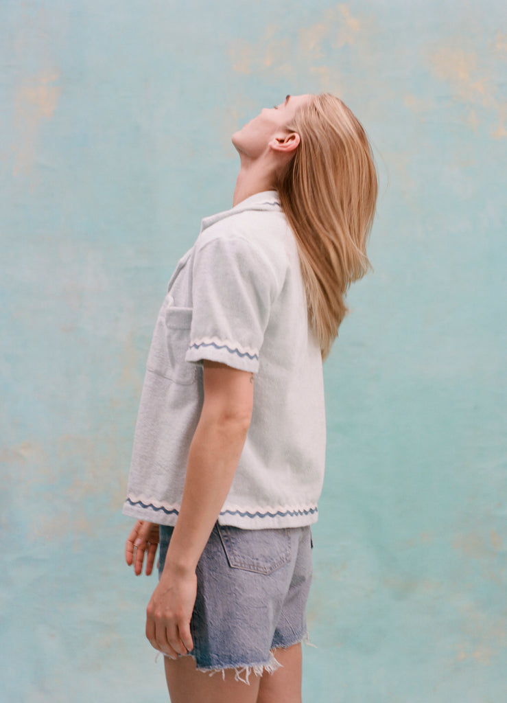 Female model with long blonde hair turned away from the camera, wearing the 'Rick Rack' Cabana ~ Crop Cut (Sky Blue) with jean shorts, shot on a light blue textured background.