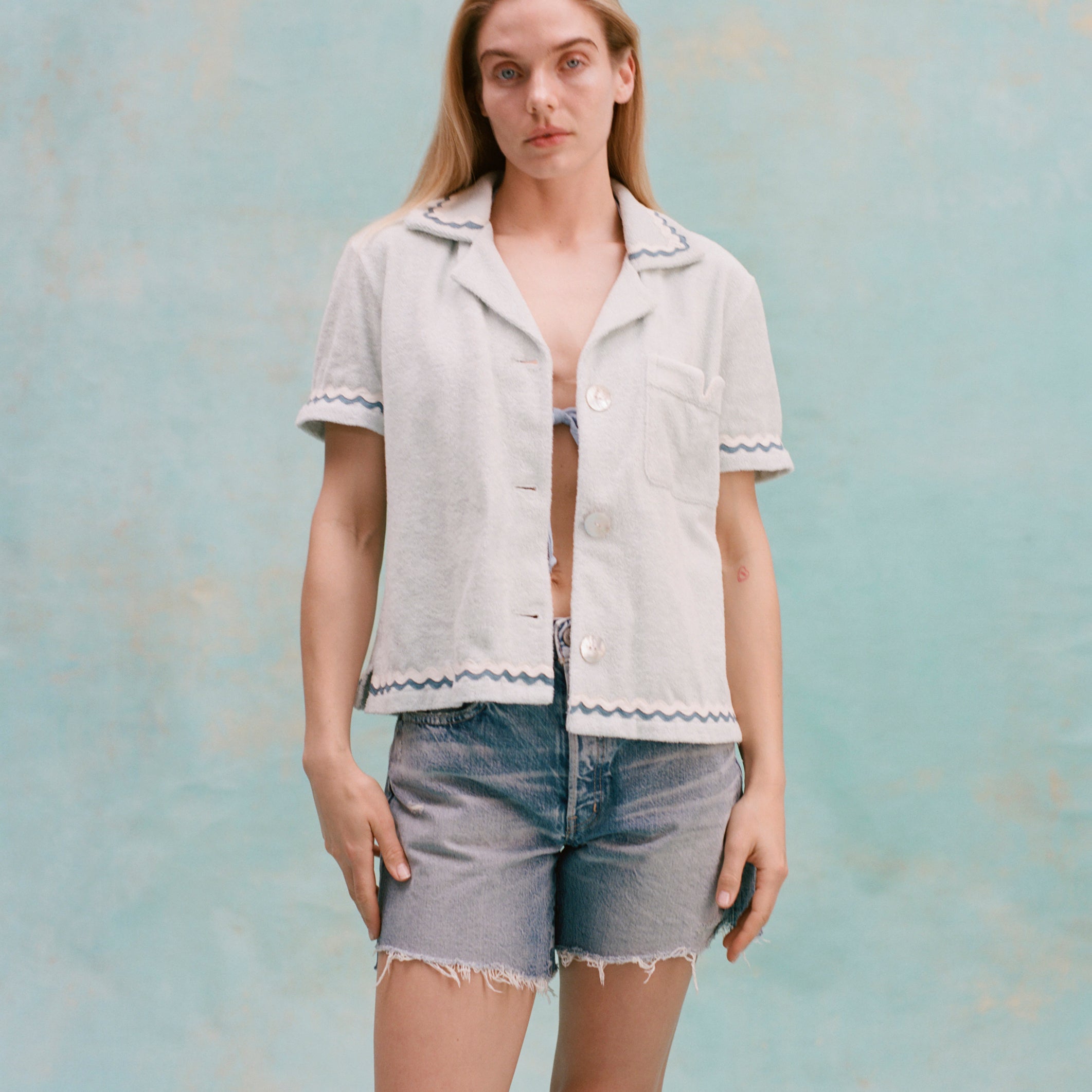 Female model with long blonde hair wearing the 'Rick Rack' Cabana ~ Crop Cut unbuttoned with teal bikini top barely visible, and jean shorts, all shot on a light blue background.