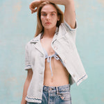 Female model with long blonde hair wearing the 'Rick Rack' ~ Cabana (Sky Blue) unbuttoned holding one hand above her head, wearing jean shorts, shot on a textured light blue background.