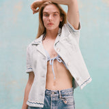 Female model with long blonde hair wearing the 'Rick Rack' ~ Cabana (Sky Blue) unbuttoned holding one hand above her head, wearing jean shorts, shot on a textured light blue background.