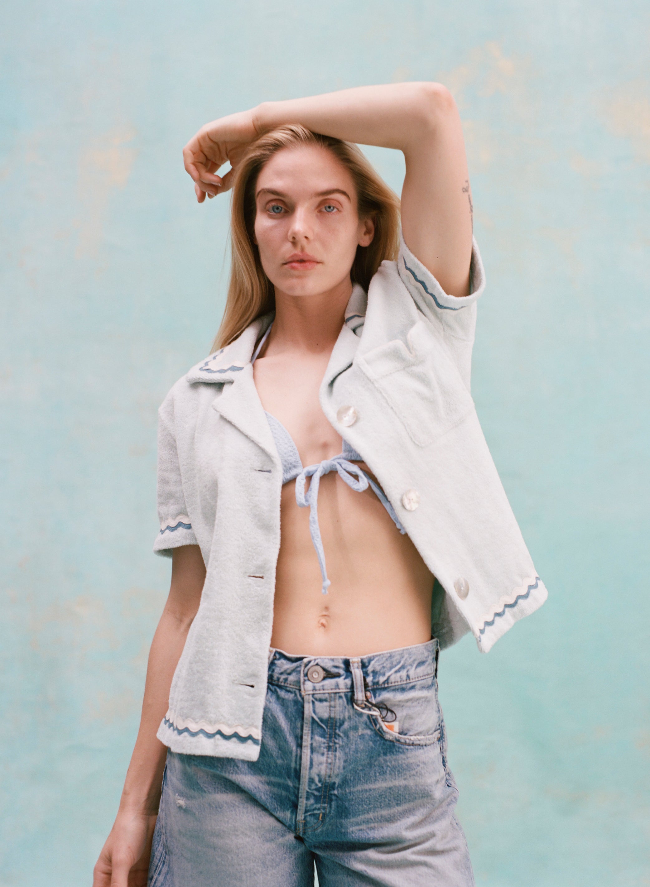 Female model with long blonde hair wearing the 'Rick Rack' ~ Cabana (Sky Blue) unbuttoned holding one hand above her head, wearing jean shorts, shot on a textured light blue background.