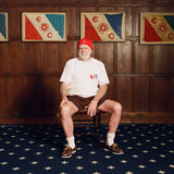 Older man seated in a wooden chair, wearing a cream terry cloth T-shirt with red stitching and a red and blue flag patch on the chest, paired with brown shorts and a red beanie featuring a small flag patch. He is posed in front of wood-paneled walls with two large flags featuring the same red and blue design and the letters “E” and “C.”