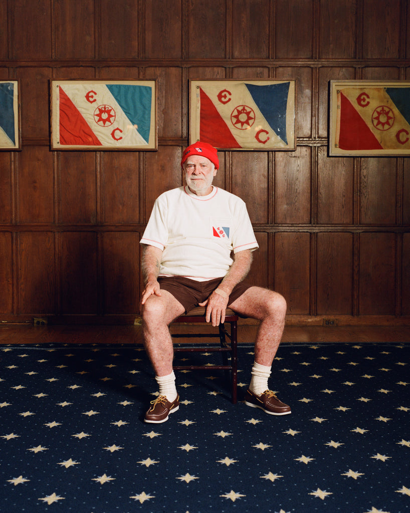 Older man seated in a wooden chair, wearing a cream terry cloth T-shirt with red stitching and a red and blue flag patch on the chest, paired with brown shorts and a red beanie featuring a small flag patch. He is posed in front of wood-paneled walls with two large flags featuring the same red and blue design and the letters “E” and “C.”