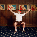 Older man seated in a chair with arms stretched wide, wearing a cream terry cloth T-shirt with red trim and a red and blue flag patch, brown shorts, a red beanie, white socks, and brown shoes. He is posed in front of a wood-paneled wall with four framed red, white, and blue flags featuring the letters “E” and “C” and a ship wheel symbol.