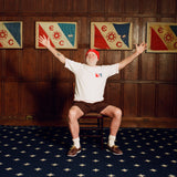 Older man seated in a chair with arms stretched wide, wearing a cream terry cloth T-shirt with red trim and a red and blue flag patch, brown shorts, a red beanie, white socks, and brown shoes. He is posed in front of a wood-paneled wall with four framed red, white, and blue flags featuring the letters “E” and “C” and a ship wheel symbol.