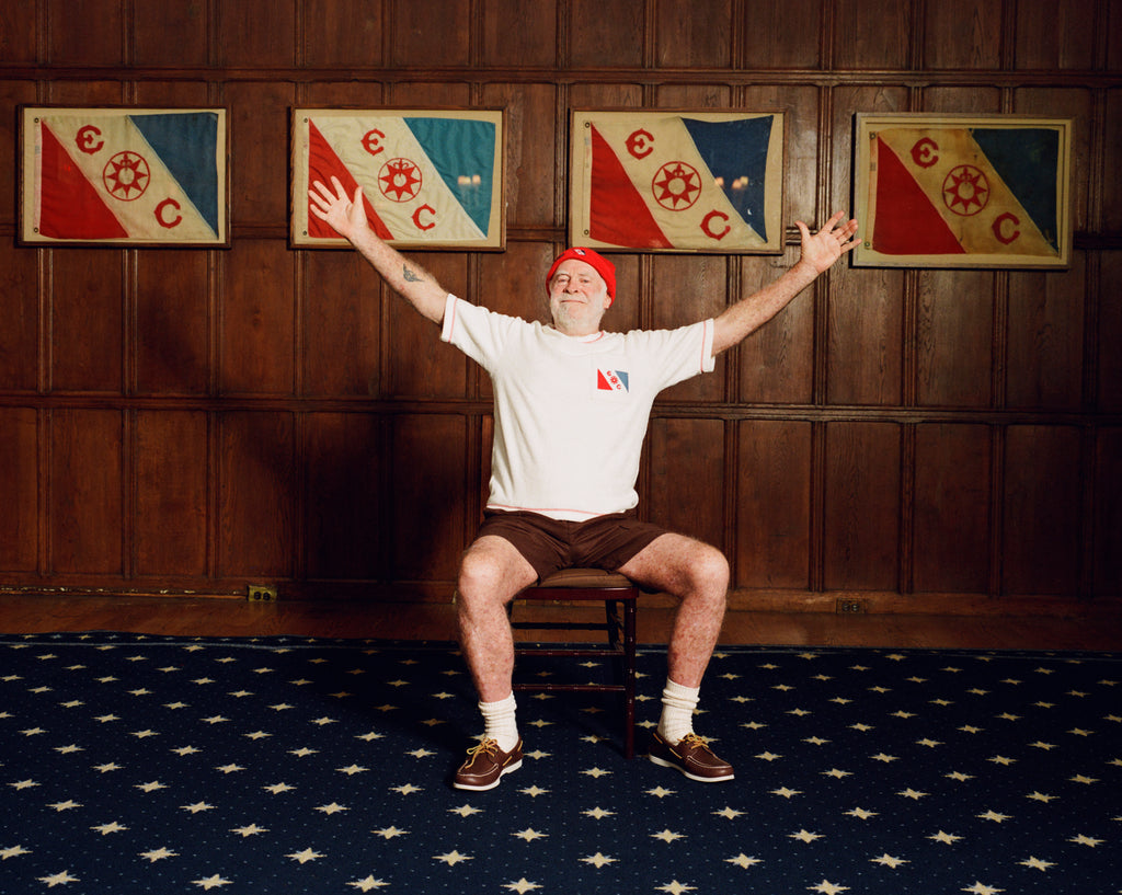 Older man seated in a chair with arms stretched wide, wearing a cream terry cloth T-shirt with red trim and a red and blue flag patch, brown shorts, a red beanie, white socks, and brown shoes. He is posed in front of a wood-paneled wall with four framed red, white, and blue flags featuring the letters “E” and “C” and a ship wheel symbol.