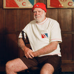 Older man seated in a wooden chair, wearing a cream terry cloth T-shirt with red stitching and a red and blue flag patch on the chest, paired with brown shorts and a red beanie featuring a small flag patch. He is posed in front of wood-paneled walls with two large flags featuring the same red and blue design and the letters “E” and “C.”