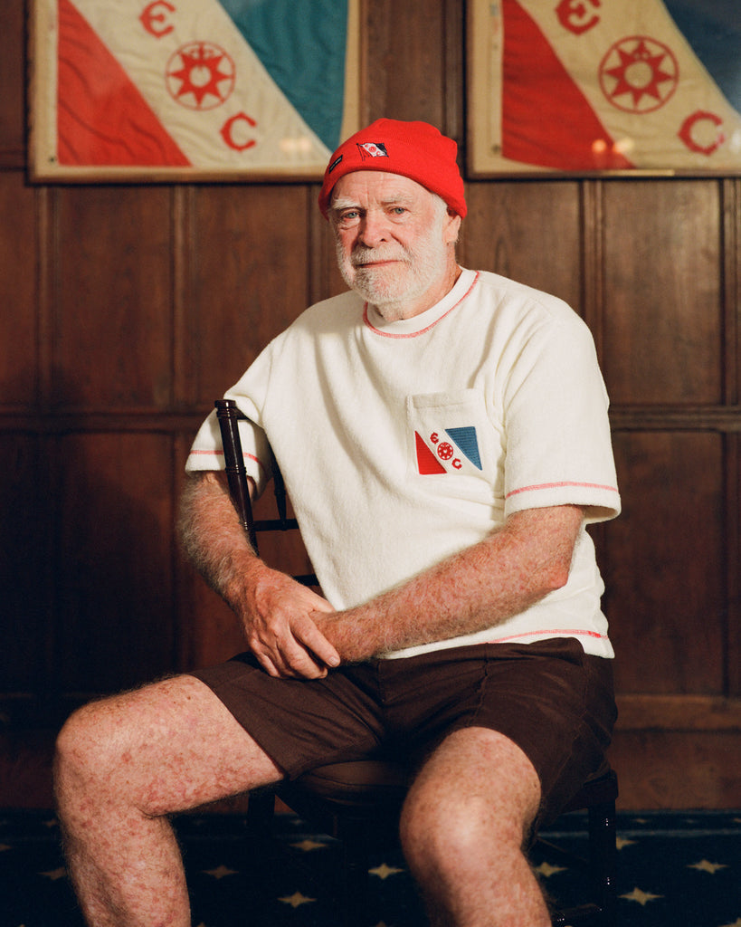 Older man seated in a wooden chair, wearing a cream terry cloth T-shirt with red stitching and a red and blue flag patch on the chest, paired with brown shorts and a red beanie featuring a small flag patch. He is posed in front of wood-paneled walls with two large flags featuring the same red and blue design and the letters “E” and “C.”