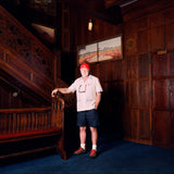 Explorer's Club Member, Brian Gaisford, is seen standing in front of a wooden staircase, wearing the Short-sleeved cream button-up shirt with the Explorer's Club Flag over the heart. 