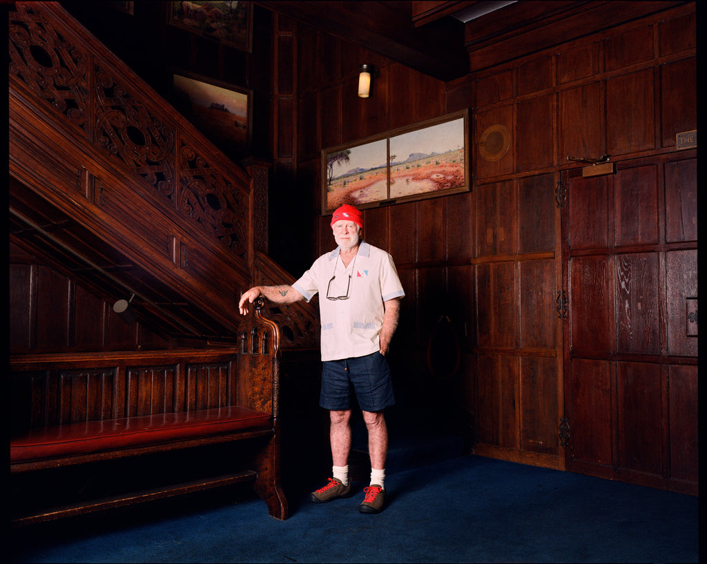 Explorer's Club Member, Brian Gaisford, is seen standing in front of a wooden staircase, wearing the Short-sleeved cream button-up shirt with the Explorer's Club Flag over the heart. 