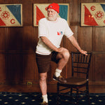 Older man standing at a wooden chair, wearing a cream terry cloth T-shirt with red stitching and a red and blue flag patch on the chest, paired with brown shorts and a red beanie featuring a small flag patch. He is posed in front of wood-paneled walls with two large flags featuring the same red and blue design and the letters “E” and “C.”