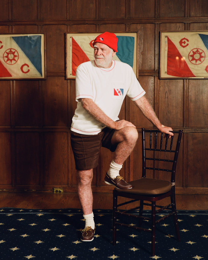 Older man standing at a wooden chair, wearing a cream terry cloth T-shirt with red stitching and a red and blue flag patch on the chest, paired with brown shorts and a red beanie featuring a small flag patch. He is posed in front of wood-paneled walls with two large flags featuring the same red and blue design and the letters “E” and “C.”