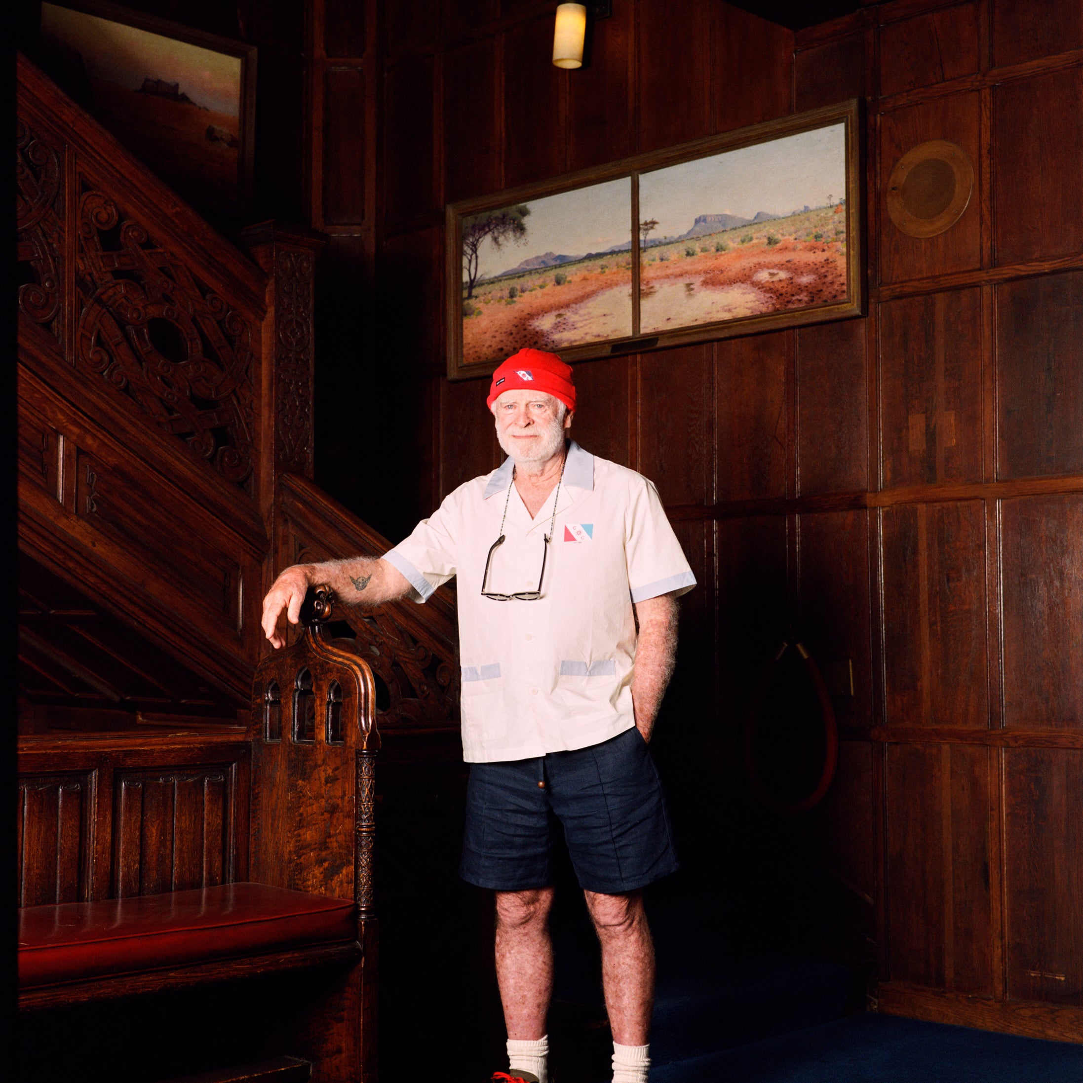 Explorer's Club Member, Brian Gaisford, is seen standing in front of a wooden staircase, wearing the Short-sleeved cream button-up shirt with the Explorer's Club Flag over the heart. 