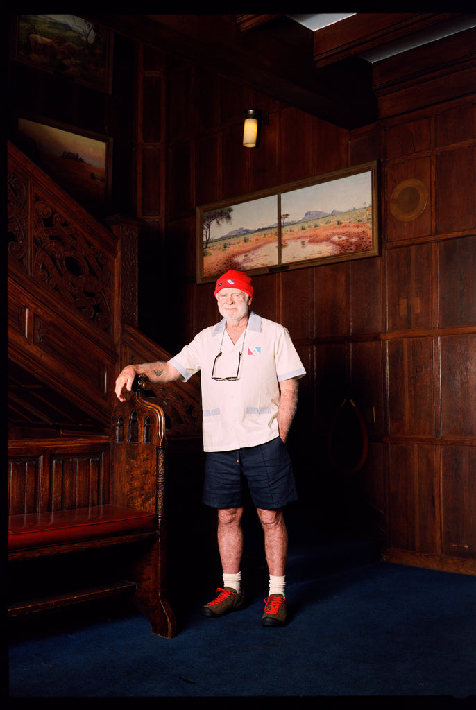 Explorer's Club Member, Brian Gaisford, is seen standing in front of a wooden staircase, wearing the Short-sleeved cream button-up shirt with the Explorer's Club Flag over the heart. 