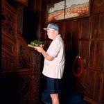 Older gentleman wearing the 'Explorer's Club' Field Uniform and our 'Explorers Club' 5-Panel Souvenir Cap. He is facing the side and carrying two books up, about to walk up the wooden stairs. 