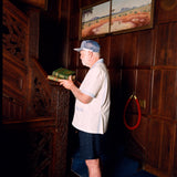 Older gentleman wearing the 'Explorer's Club' Field Uniform and our 'Explorers Club' 5-Panel Souvenir Cap. He is facing the side and carrying two books up, about to walk up the wooden stairs. 