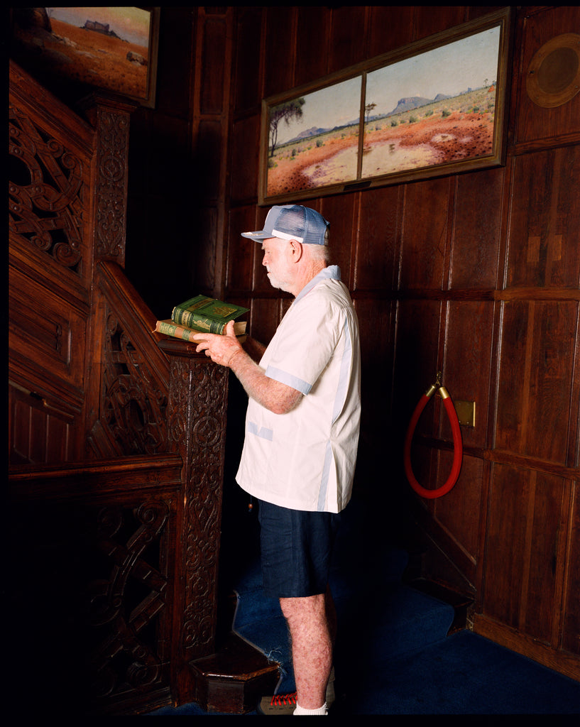 Older gentleman wearing the 'Explorer's Club' Field Uniform and our 'Explorers Club' 5-Panel Souvenir Cap. He is facing the side and carrying two books up, about to walk up the wooden stairs. 
