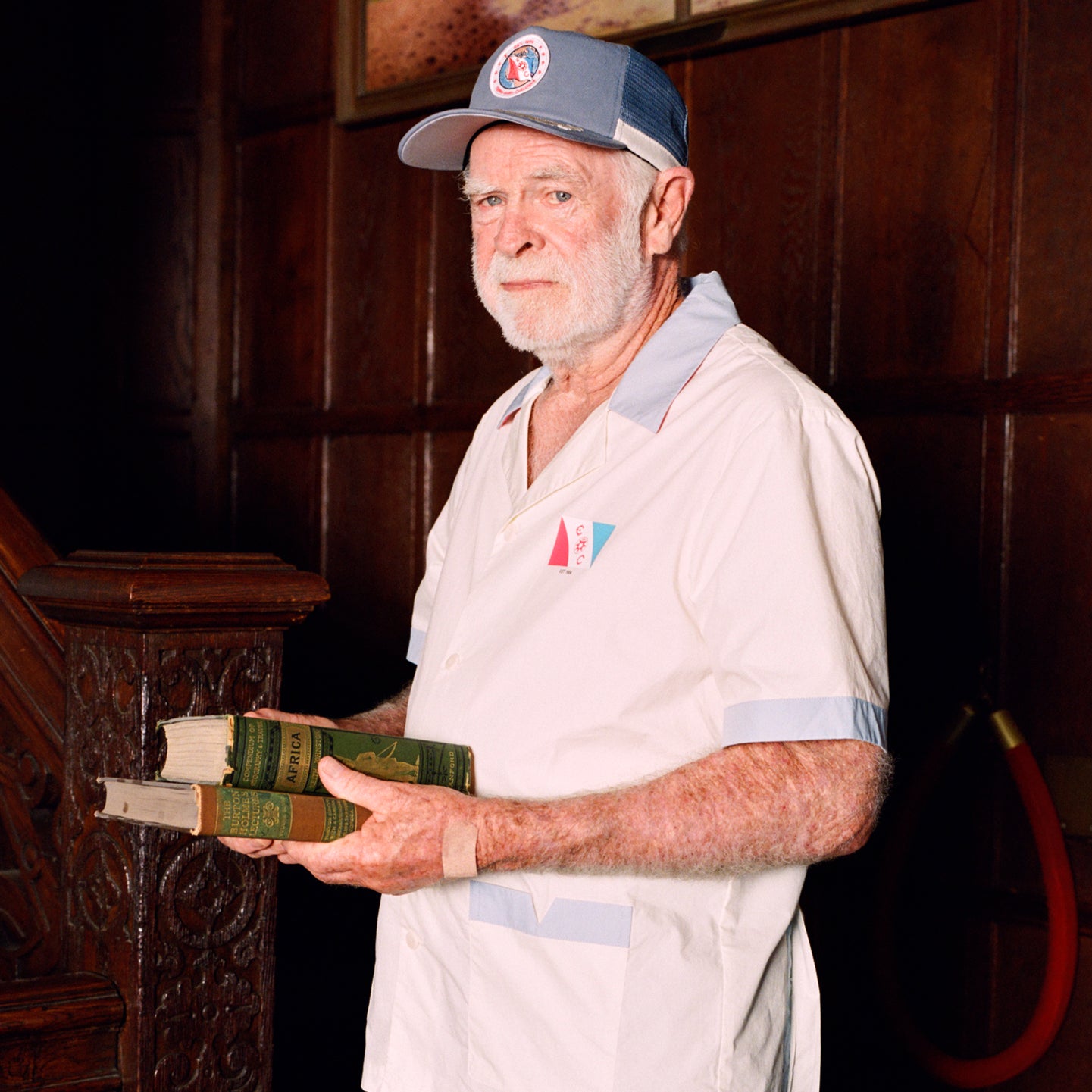Older gentleman wearing the 'Explorer's Club' Field Uniform and our 'Explorers Club' 5-Panel Souvenir Cap. He is facing the side and carrying two books.