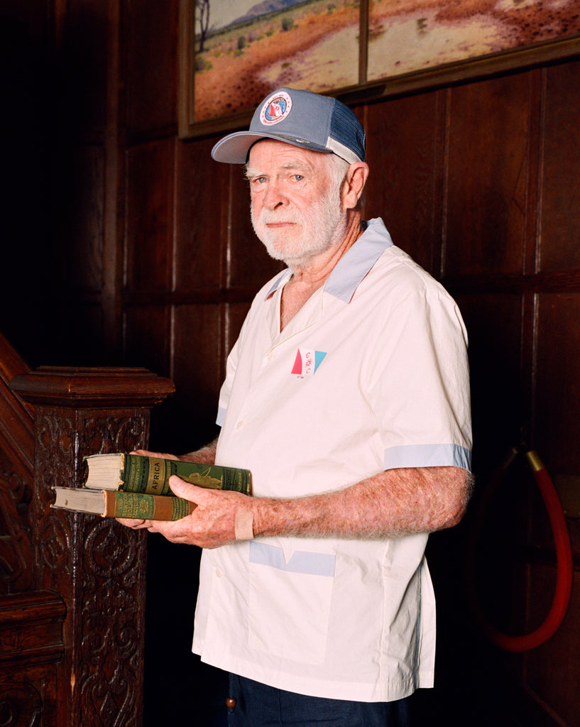 Older gentleman wearing the 'Explorer's Club' Field Uniform and our 'Explorers Club' 5-Panel Souvenir Cap. He is facing the side and carrying two books.