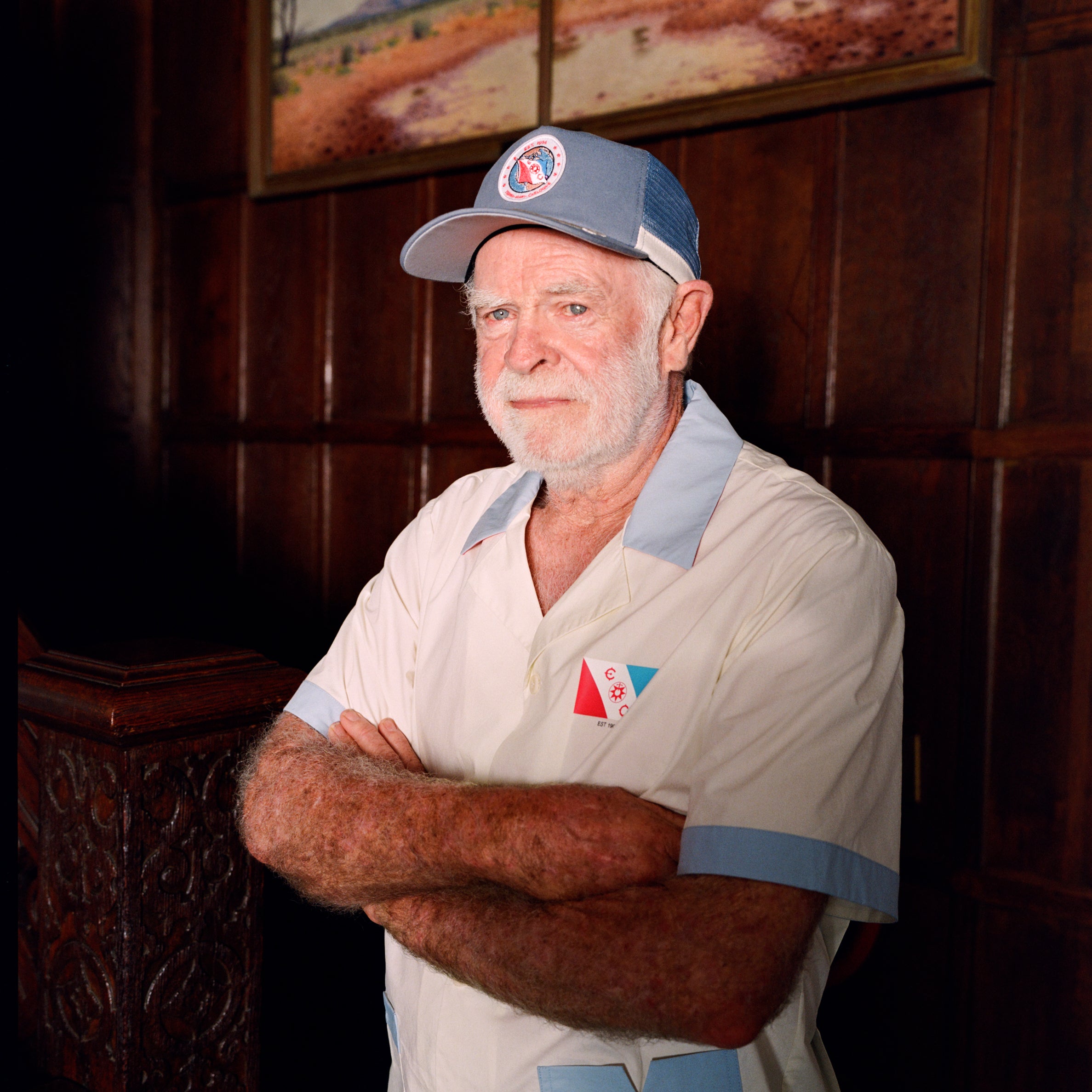 Older gentleman wearing the 'Explorer's Club' Field Uniform and our 'Explorers Club' 5-Panel Souvenir Cap. His arms are crossed. 