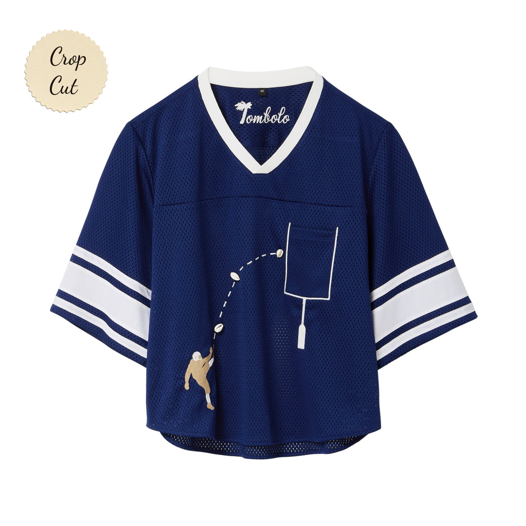 Navy blue mesh crop-cut shirt with white stripes on the sleeves, a white V-neck, and an embroidered football design showing a player kicking a football toward a goalpost outlined around the chest pocket.