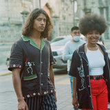 Male model wearing 'Picante' cabana shirt walking in the street, accompanied by a female model wearing a different style. 