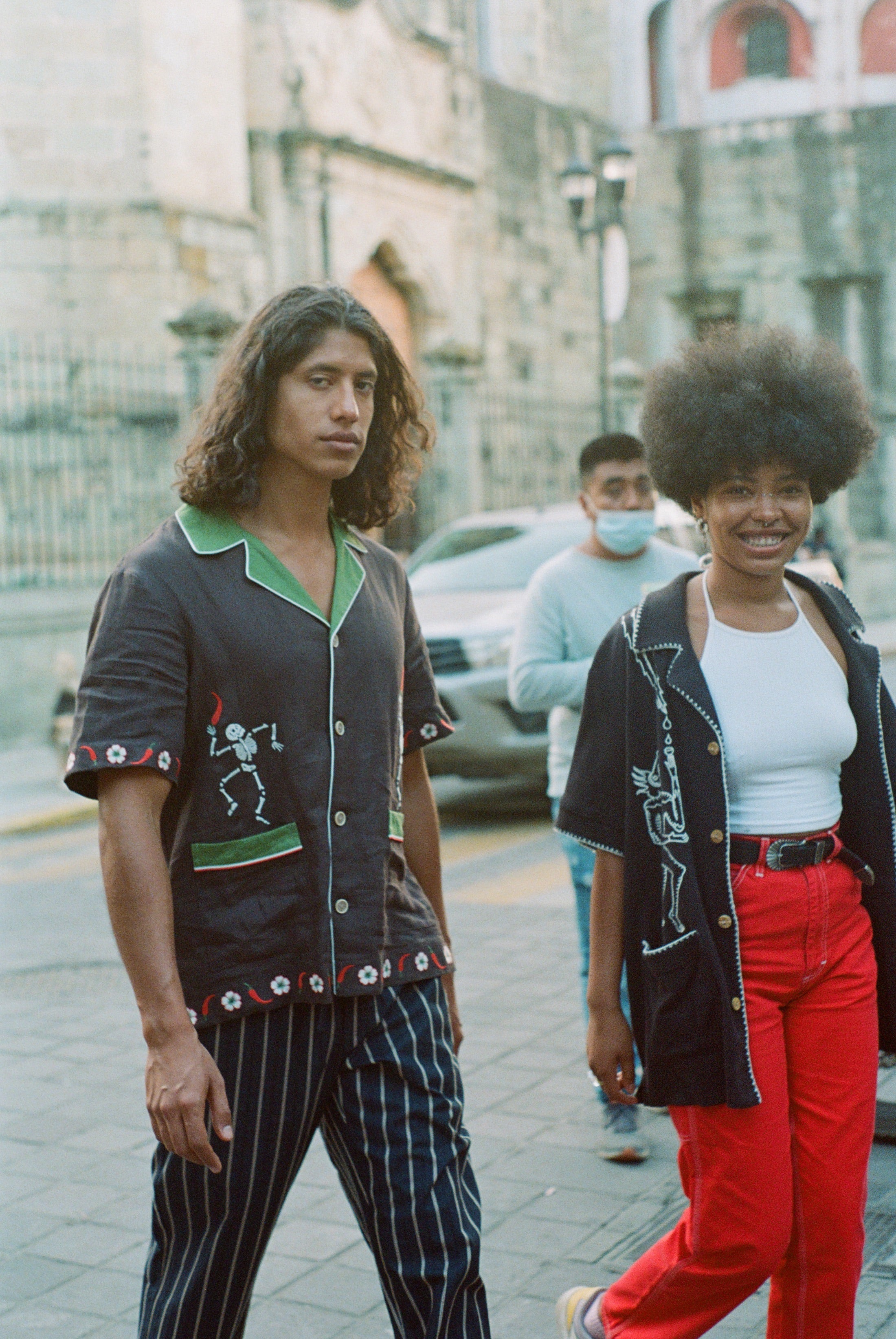 Male model wearing 'Picante' cabana shirt walking in the street, accompanied by a female model wearing a different style. 