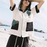 Female model in sunglasses wearing full linen 'Tombarolo' Cabana Set Casual, linen cabana style in pale yellow with eggplant color collar, short sleeves and pockets, Appliqué and edge-embroidered grapes adorn one pocket and the wearer's left chest, Eggplant colored shorts with grapes embroidered on one leg