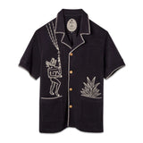 A black short-sleeve terry cloth shirt with light tan embroidery. On the left chest is a stylized figure with exaggerated features, standing beneath lightning bolts and raindrops. On the right pocket is an embroidered agave plant. The shirt has wooden buttons, front patch pockets, and contrast whipstitch detailing along the collar, cuffs, and hems. ('Agua Magica' Cabana)