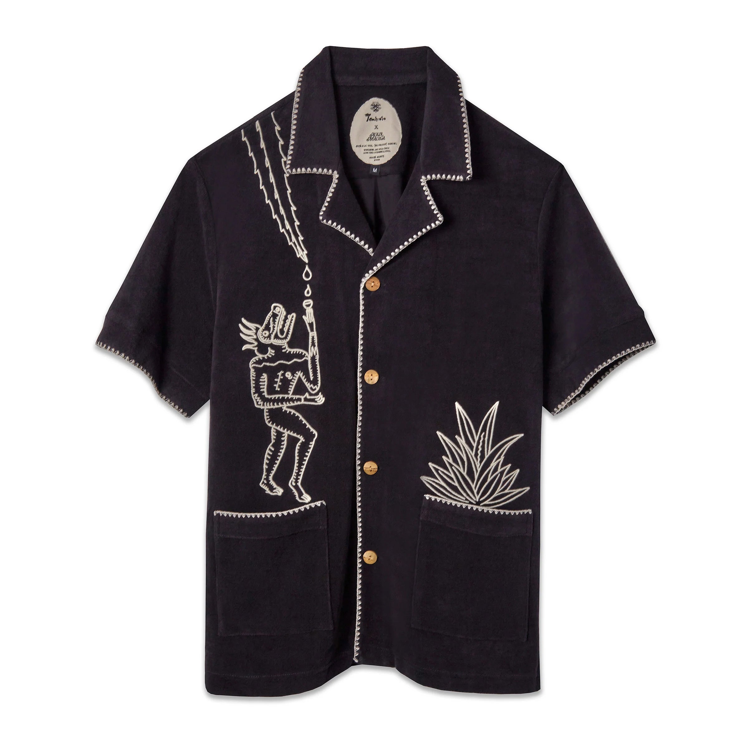 A black short-sleeve terry cloth shirt with light tan embroidery. On the left chest is a stylized figure with exaggerated features, standing beneath lightning bolts and raindrops. On the right pocket is an embroidered agave plant. The shirt has wooden buttons, front patch pockets, and contrast whipstitch detailing along the collar, cuffs, and hems. ('Agua Magica' Cabana)